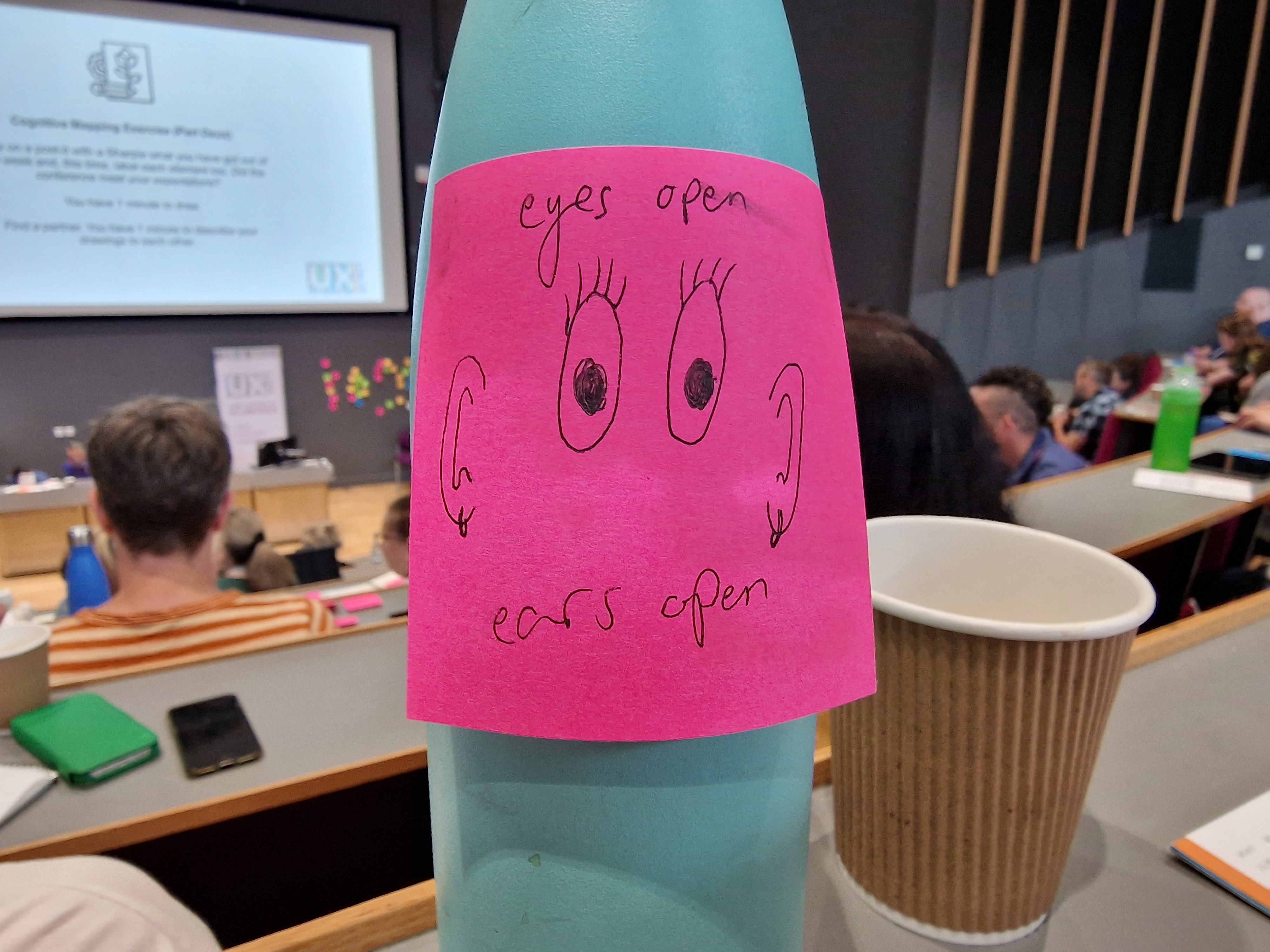 UXLibs 2023 Mint Green Water Bottle with a Pink Post it note with a drawing of 2 eyes and 2 ears with the text Eyes Open Ears Open